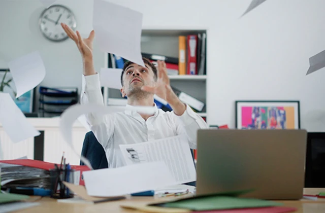 A person sits at a cluttered desk, surrounded by piles of paper. He looks overwhelmed and are throwing their hands up in the air.