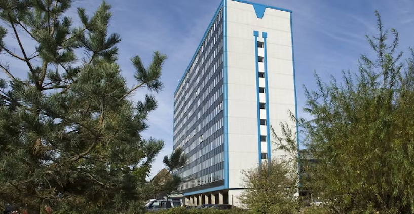 A tall, modern office building with blue vertical accents, surrounded by green trees under a clear sky.