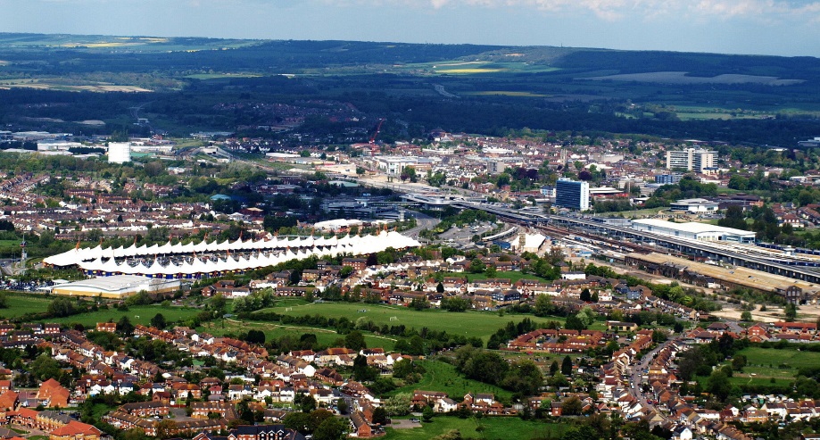 Aerial view of a town with residential neighborhoods, a large white-roofed building, railway tracks, and open green landscape stretching into the distance.