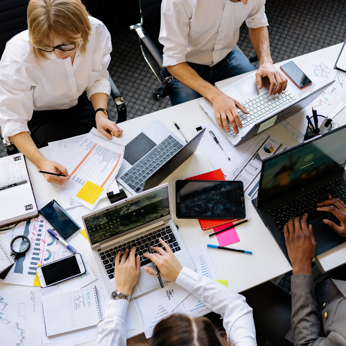 Aerial view of a busy office desk with four people working on laptops and writing. Papers, sticky notes, and devices are scattered, indicating a collaborative, focused workspace.