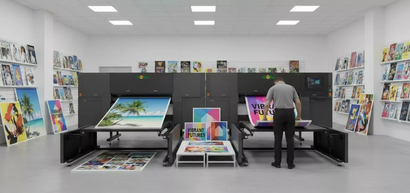 A person stands behind Flora X20UV Ricoh Printer, looking at the vibrant image of a tropical beach scene being printed. The printer is a large, black machine with multiple sections, and another similar machine is next to it, also printing a colorful design. On the wall behind the printers, a rack of printed materials is displayed. In the foreground, there are more printed graphics, including some with geometric patterns and the text "Vibrant Futures".