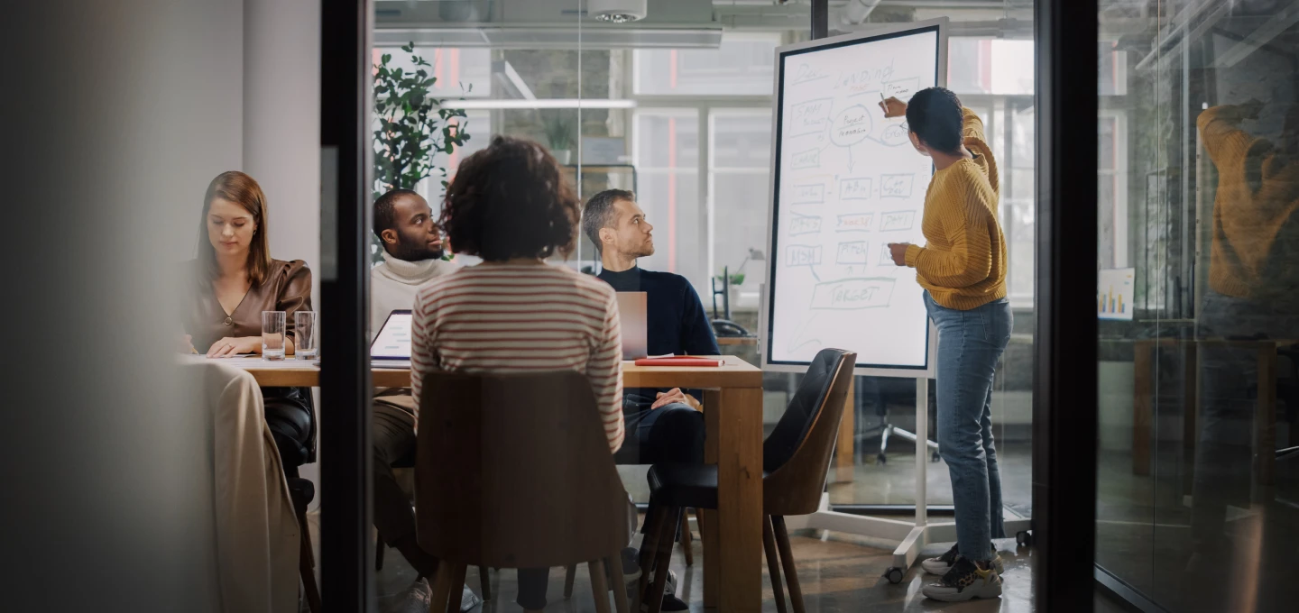 A person presents a diagram on a flip chart while four people sit around a table, listening attentively in a modern office setting