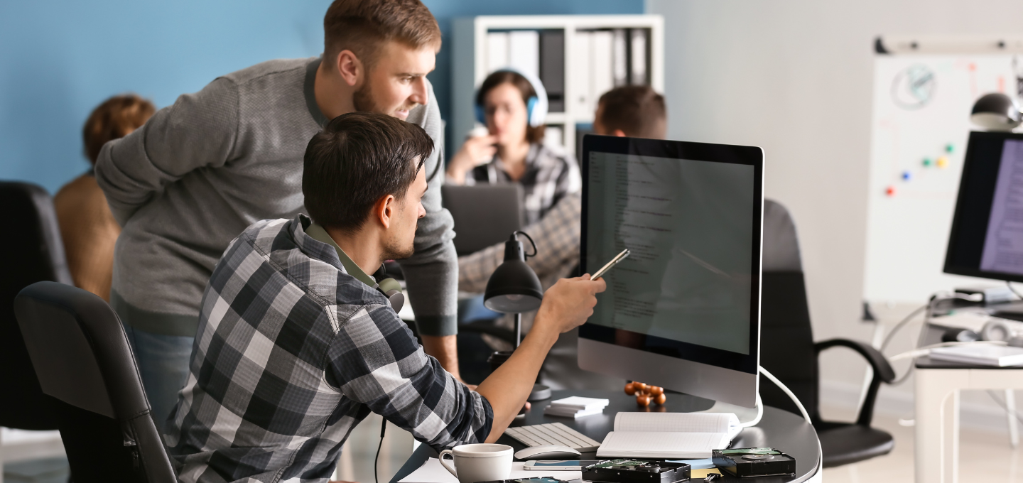Two men in a modern office are focused on a computer screen. One points with a pen, suggesting teamwork. Others work in the background, creating a collaborative atmosphere.