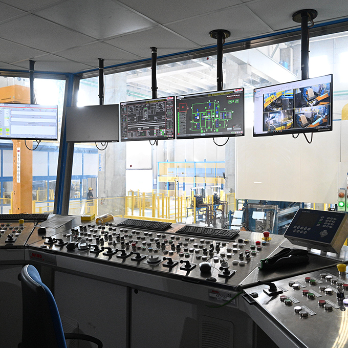 Operator control room with multiple monitors displaying technical schematics, real-time industrial data, and CCTV feeds, overlooking a metal processing or rolling mill floor. Features a complex console with numerous buttons and levers for machinery operation in a manufacturing plant of Laminazione Sottile