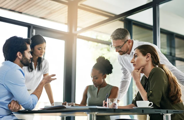 A diverse group of five professionals engaged in a discussion around a table in a modern, well-lit office.