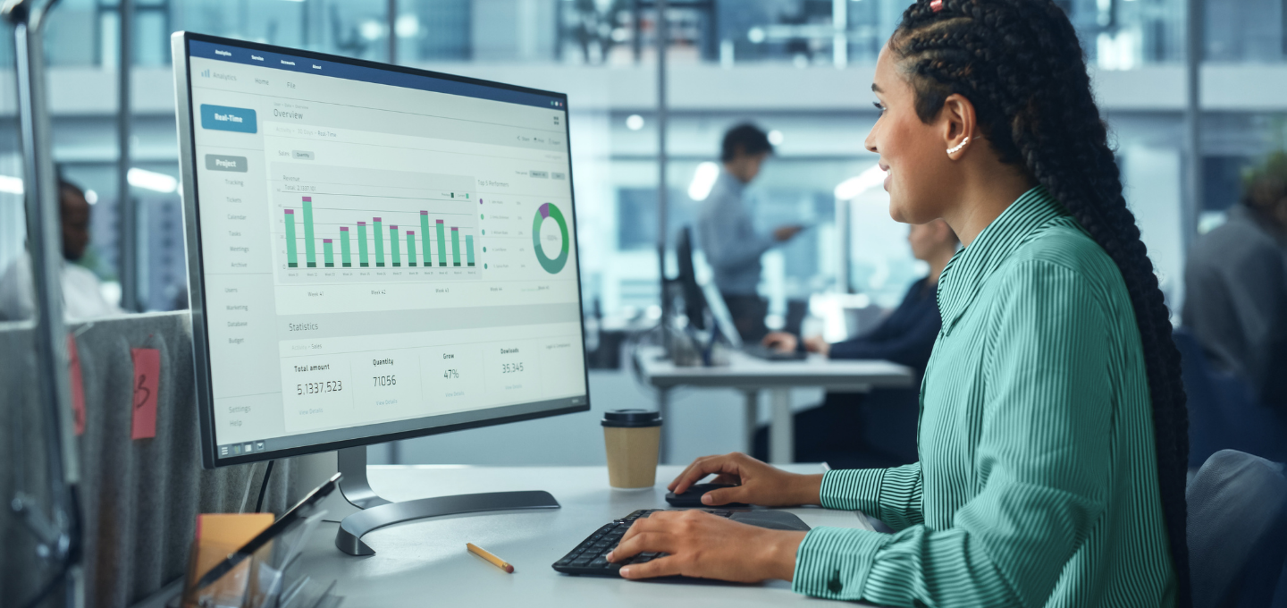 A woman in a green blouse works at a desk in an office, analyzing graphs on a monitor. The setting conveys productivity and focus.