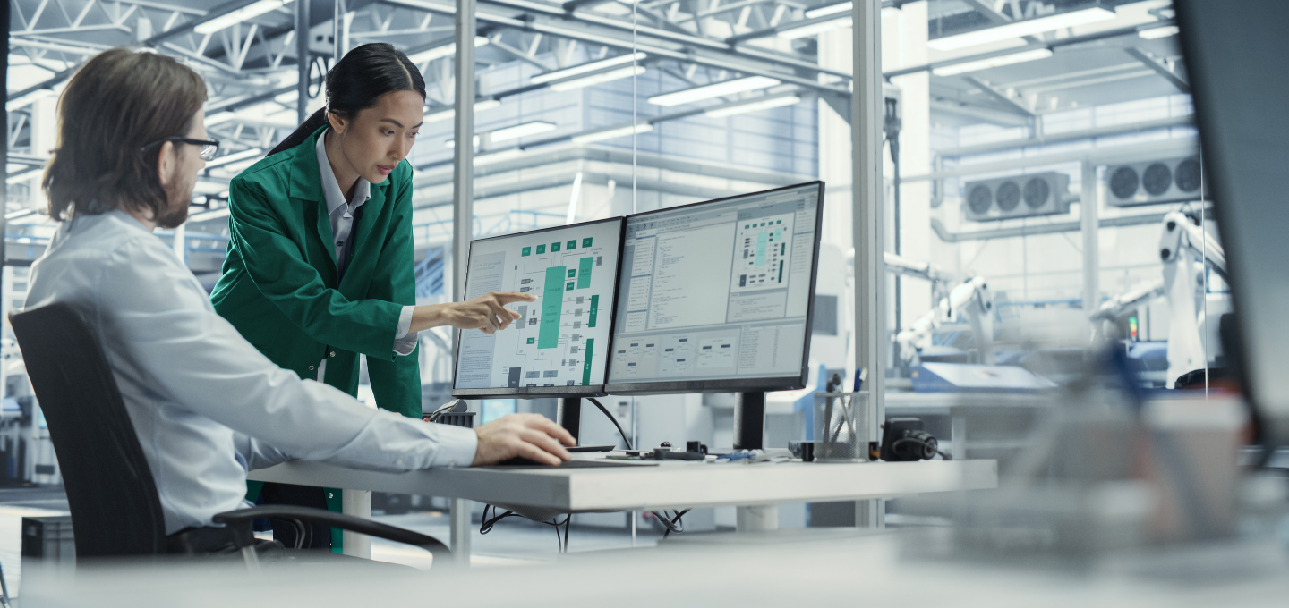 Two engineers in a modern factory review data on dual monitors. One, seated, watches as the other, standing, points at the screens. High-tech, collaborative.